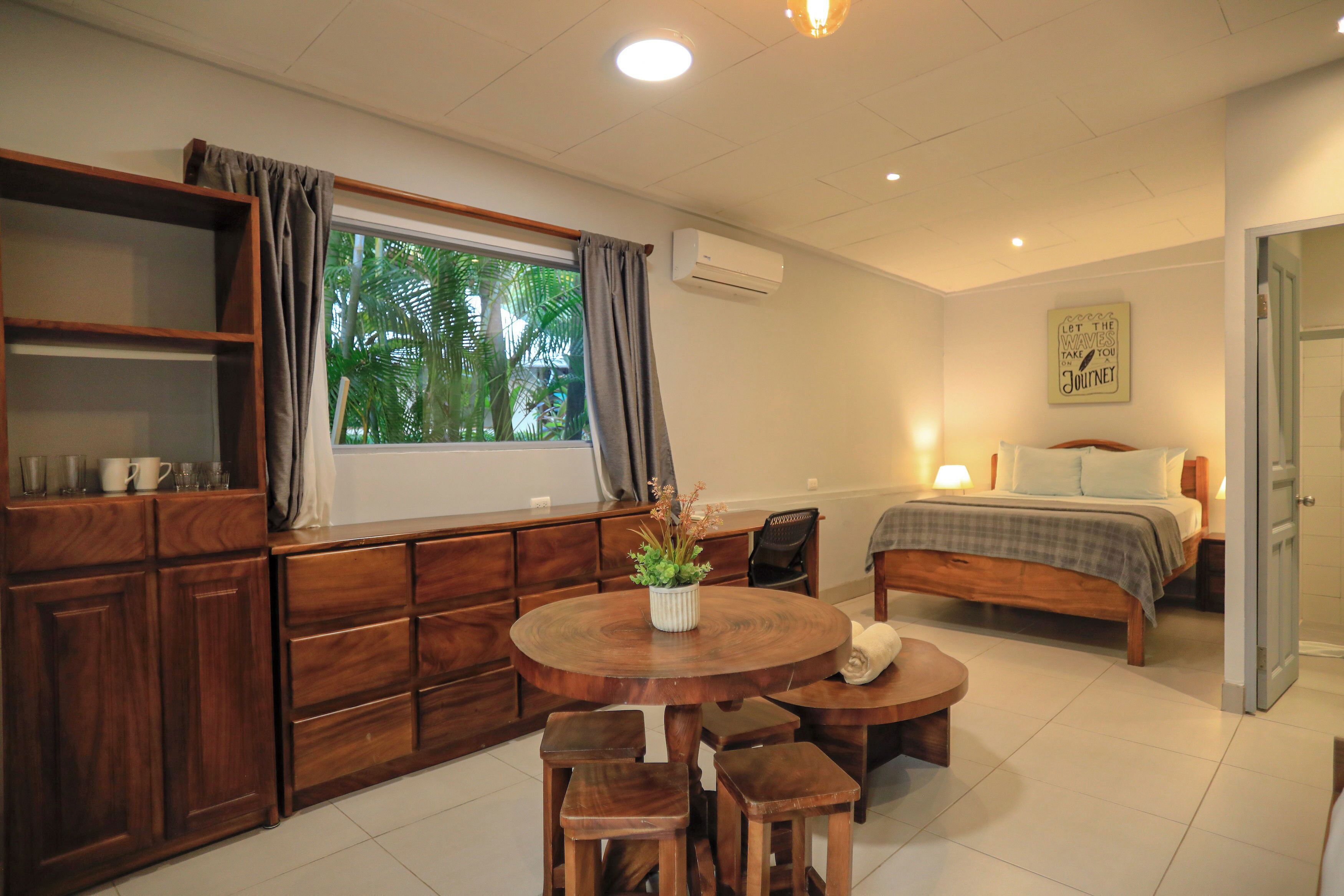 A bedroom with a bed, bedside tables, a window with curtains, a bedside lamp, and a wall-mounted air conditioner.