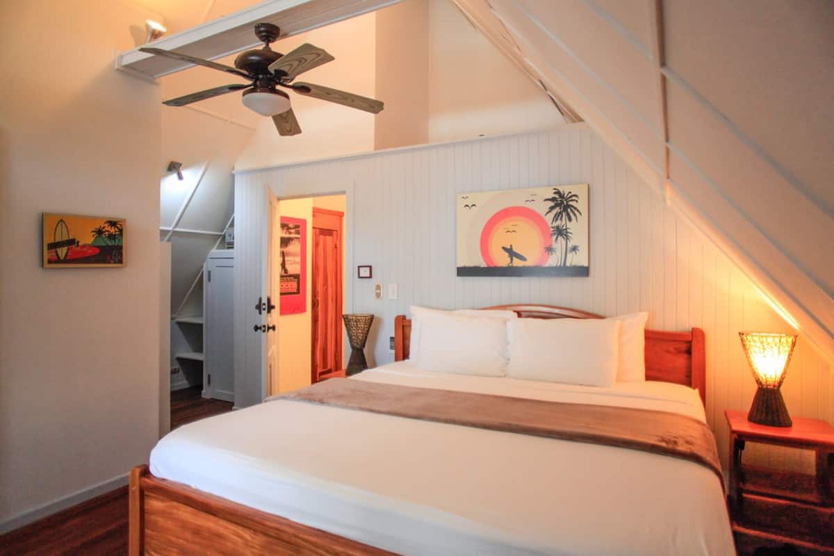 A bedroom with a bed, a ceiling fan, a nightstand with a lamp, and a painting on the wall.