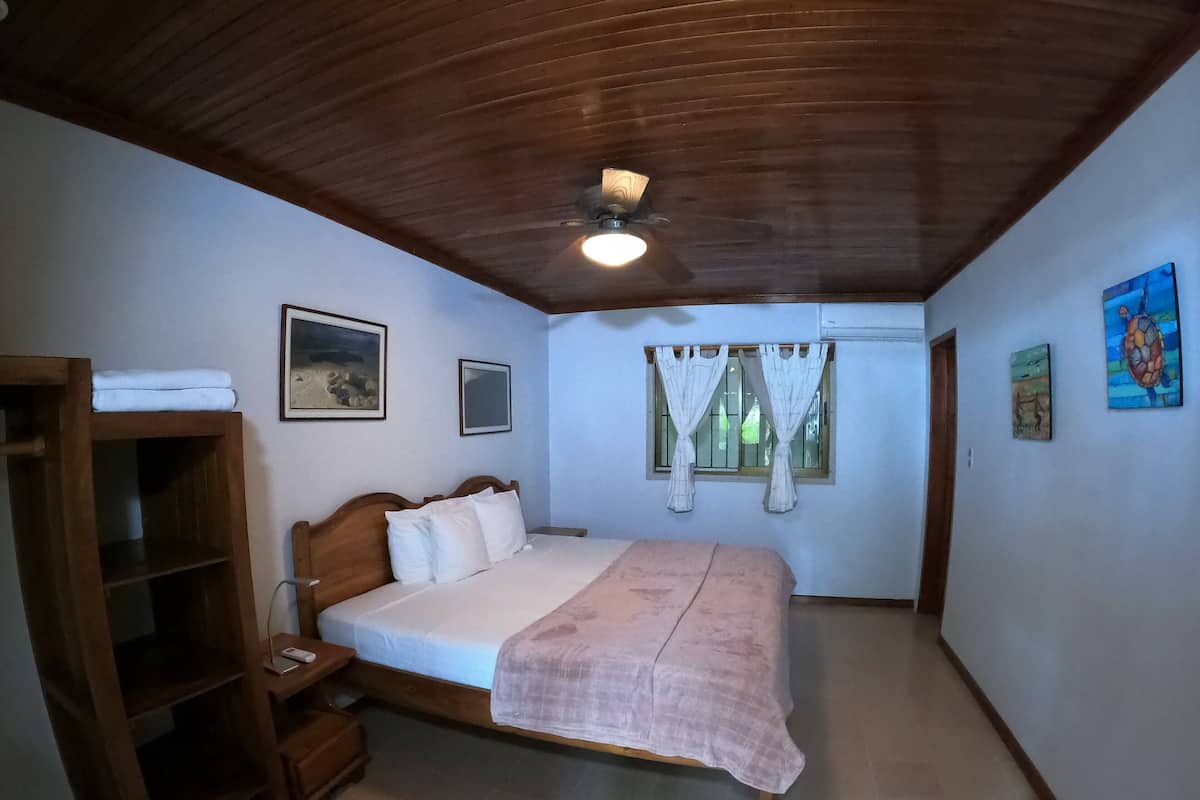 A bedroom with a wooden ceiling, a bed with white bedding, a wooden wardrobe, and two windows with curtains.