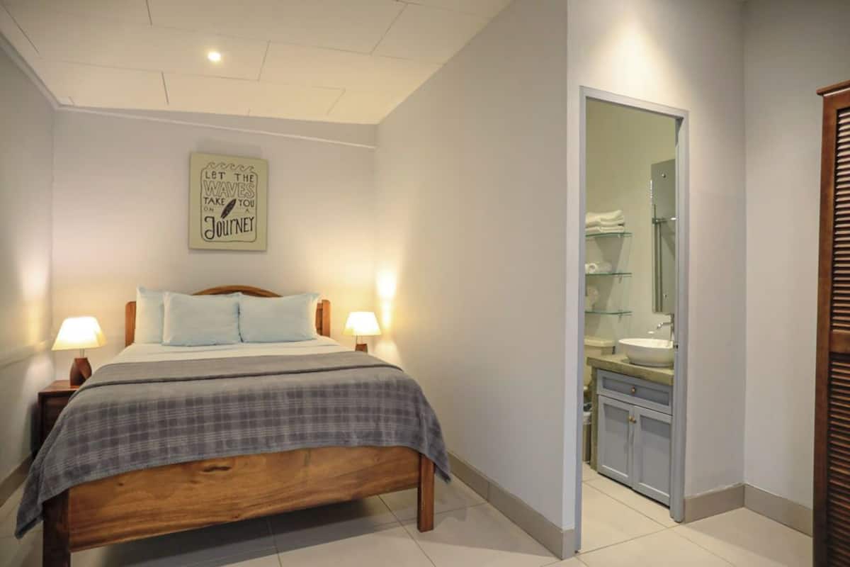 A bedroom with a bed, bedside lamps, a wall-mounted sign, a bathroom with a sink and mirror.