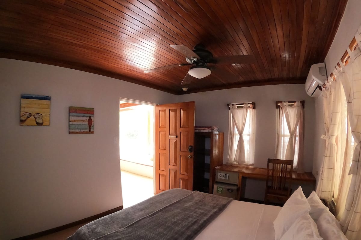 A bedroom with a wooden ceiling, a bed with white pillows, a desk with a chair, and a window with curtains.