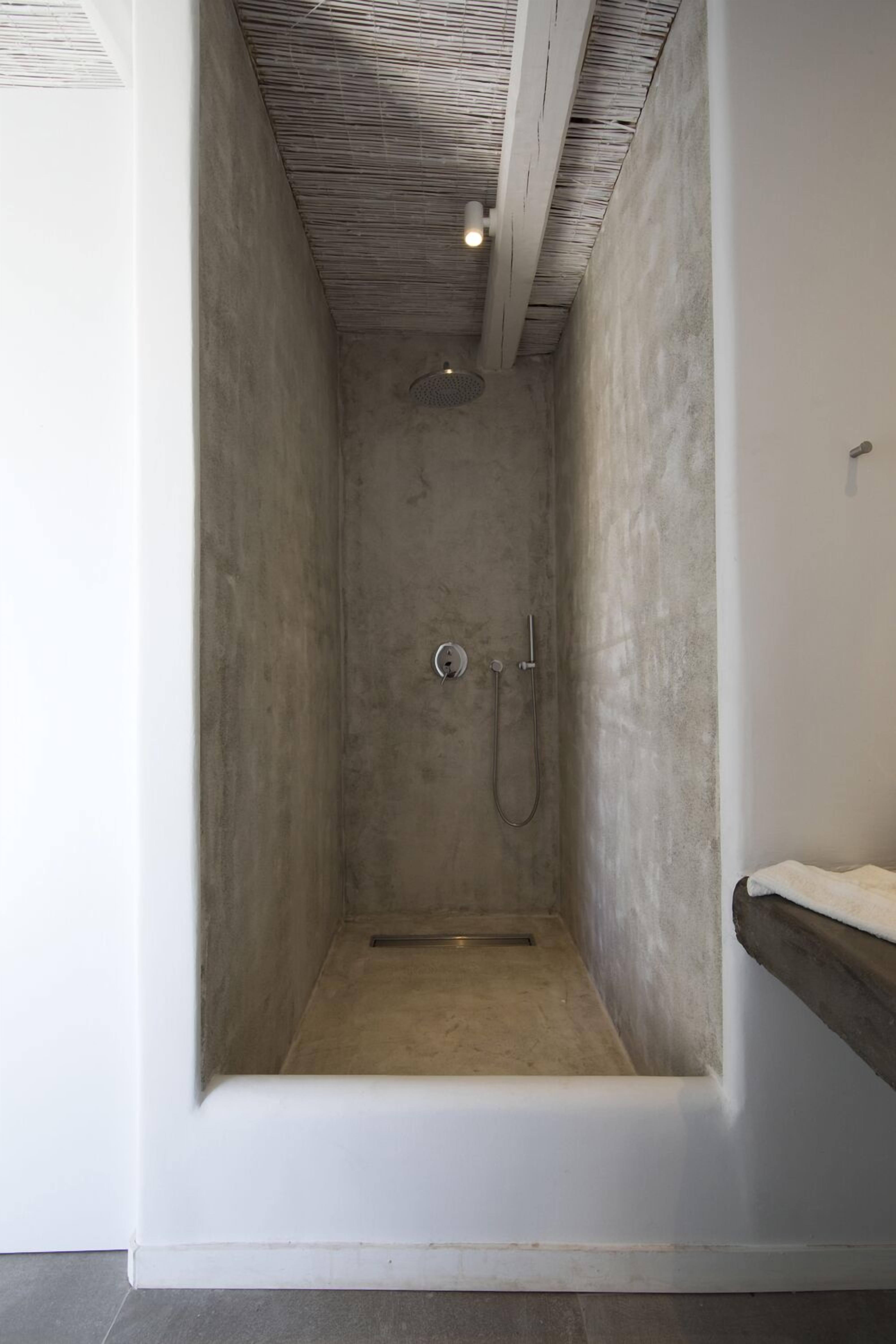 A narrow shower stall with a concrete wall and ceiling, a showerhead, and a small shelf with a towel.