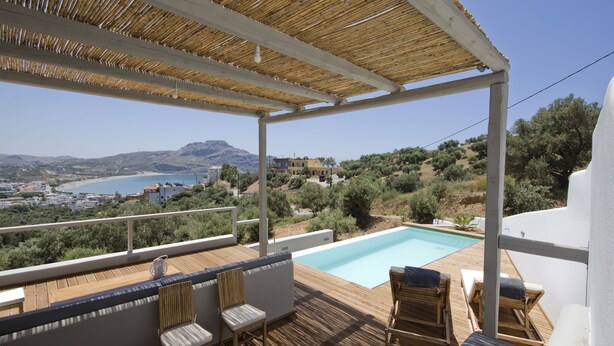 A wooden deck with a pool, outdoor seating, and a view of the sea and hills. - Villa Nissos (Agios Vasileios)