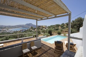 A wooden deck with a pool, outdoor seating, and a view of the sea and hills. - Villa Nissos (Agios Vasileios)