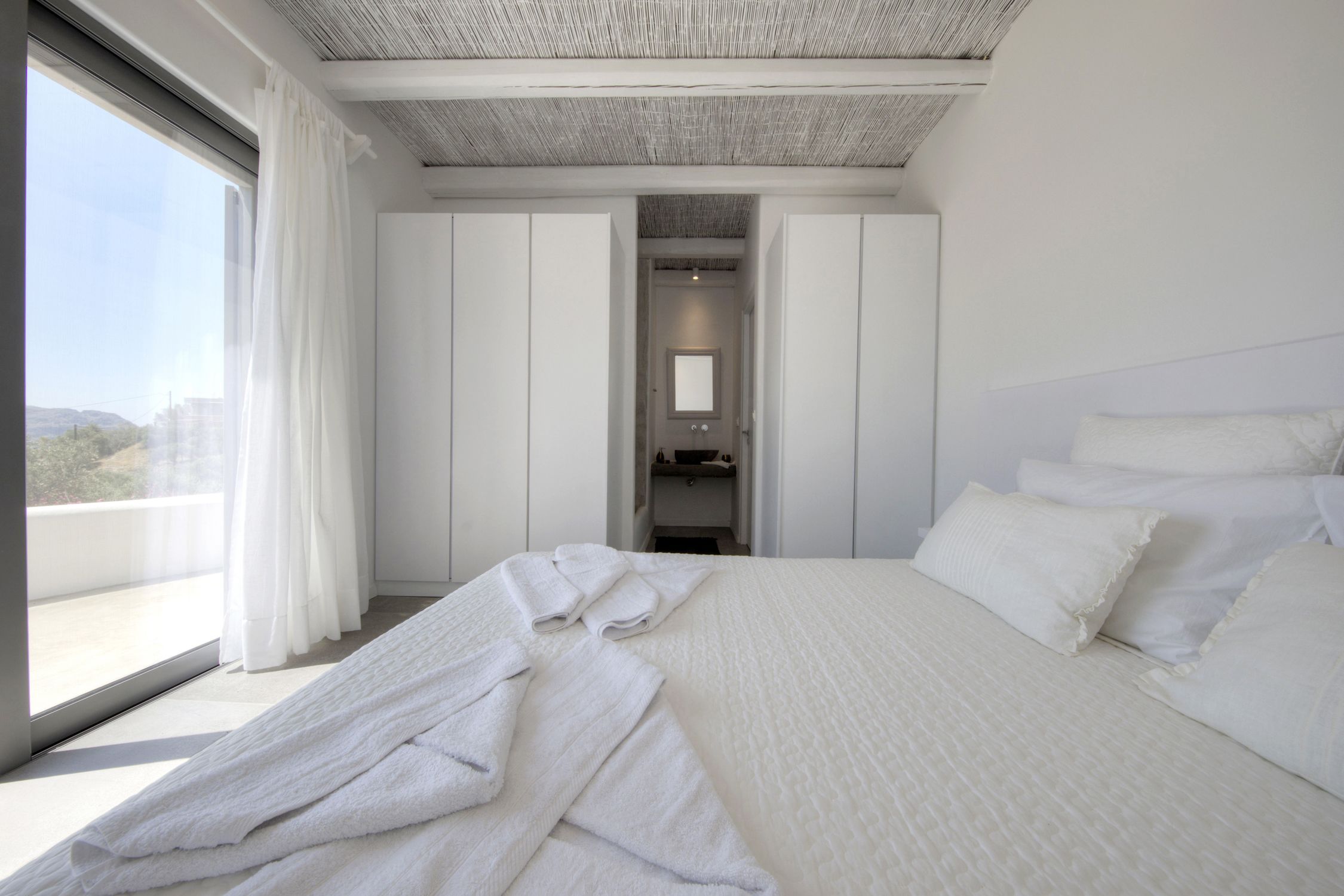 A modern bedroom with a large bed, white linens, and a view of the city through a large window.