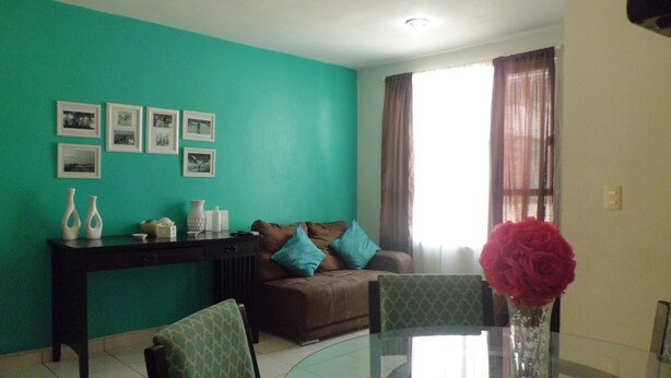 A living room with teal walls, a glass dining table, a sofa with blue cushions, and a vase with red flowers. - Condo Maple (Mazatlán)