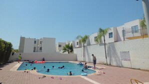 A swimming pool area with multiple people swimming and playing, surrounded by a white wall and palm trees. - Condo Maple (Mazatlán)
