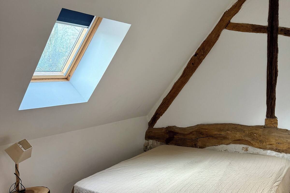 A bedroom with a sloped ceiling, a skylight, a bed with a quilted cover, a wooden nightstand, and a lamp.