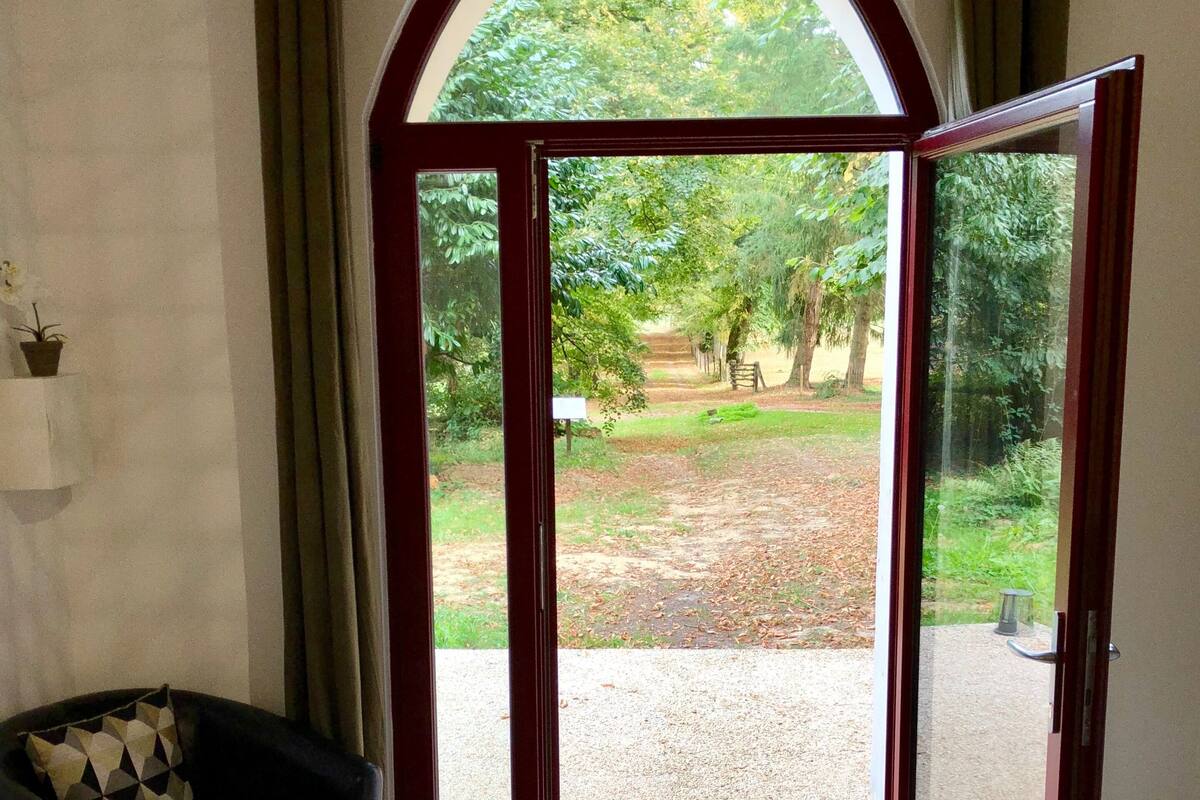 A room with a black leather sofa, a wooden coffee table, and a large arched glass door leading to an outdoor area with a gravel path and greenery.