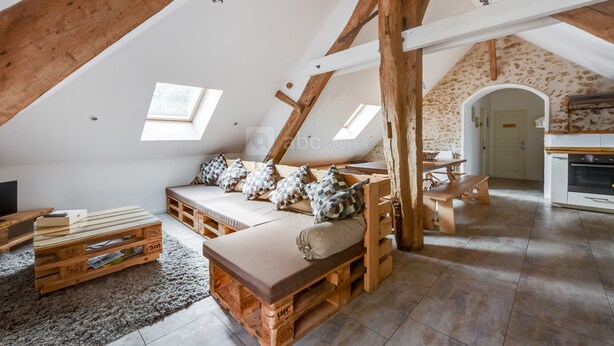 A loft-style room with a sloped ceiling, wooden beams, a built-in bench with cushions, a small table, and a kitchen area. - Domaine de la Geneste (Châteaufort)