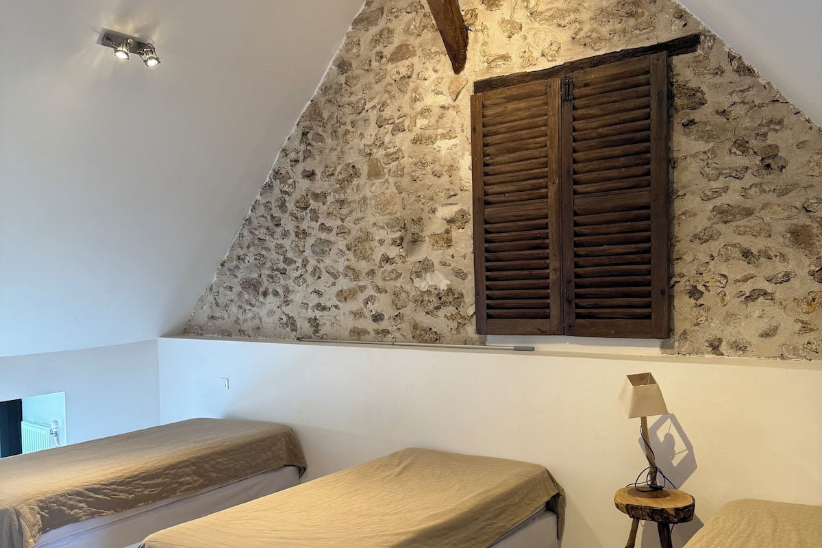 A bedroom with two single beds, a wooden beam ceiling, a stone wall, and a window with shutters.