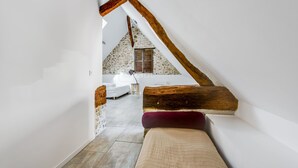 A narrow attic bedroom with exposed wooden beams, a bed with a red pillow, and a view of a bathroom with a white bathtub. - Domaine de la Geneste (Châteaufort)