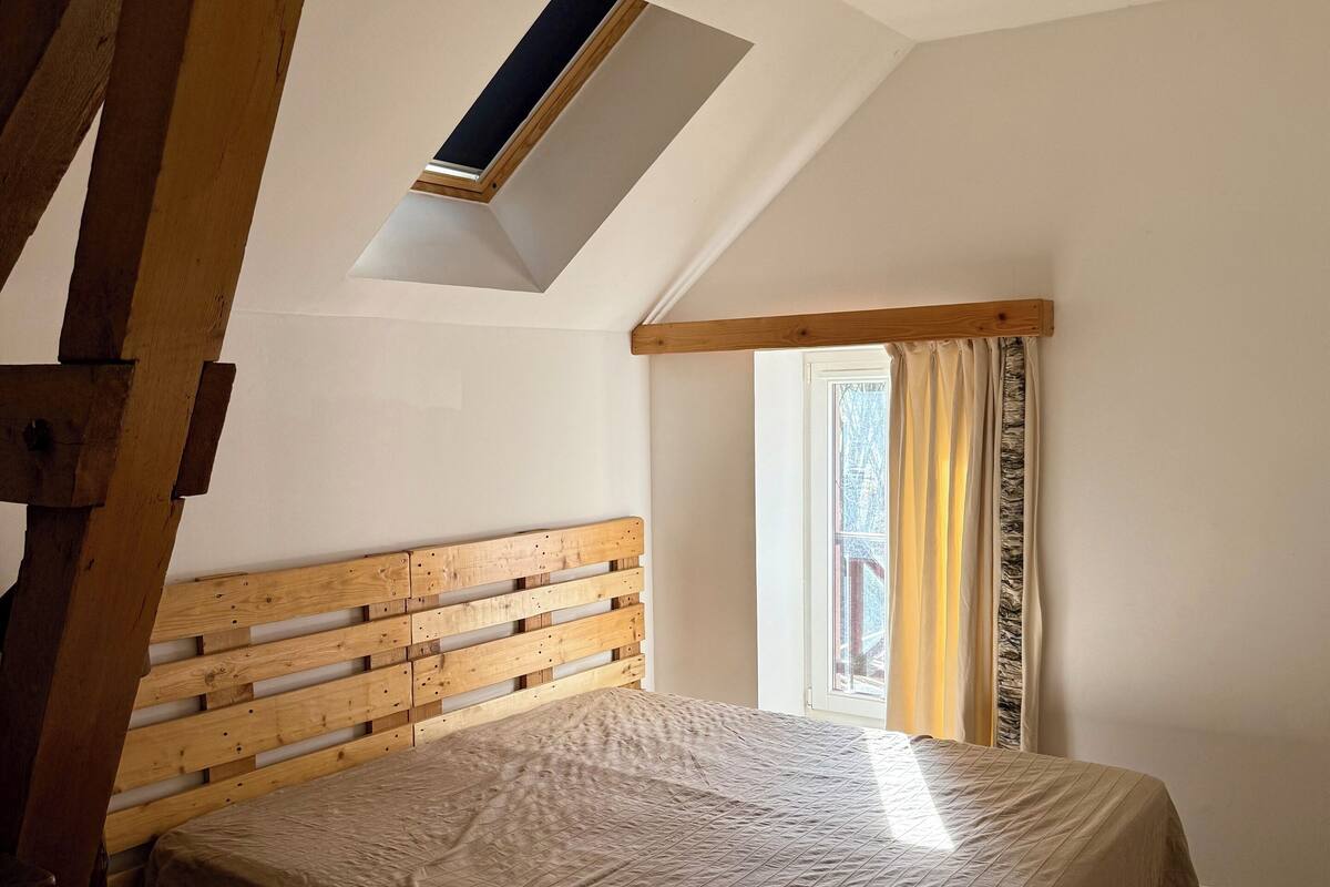 A bedroom with a sloped ceiling, a wooden headboard, a bed with a beige bedspread, a skylight, and a window with curtains.