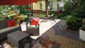 An outdoor seating area with wicker furniture and a colorful rug, surrounded by lush greenery and flowers. - OW Sargus (Wladyslawowo)