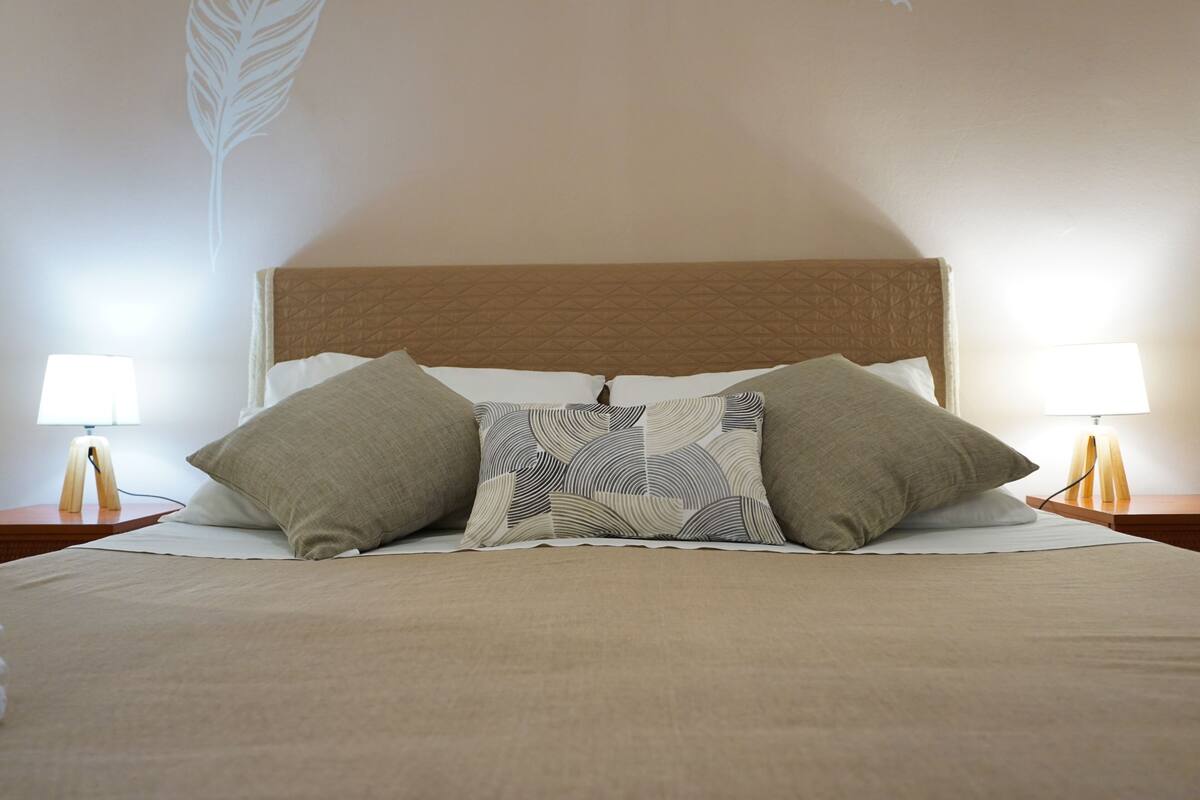 A neatly made bed with a patterned pillow, two bedside lamps, and a wall with a leaf design.