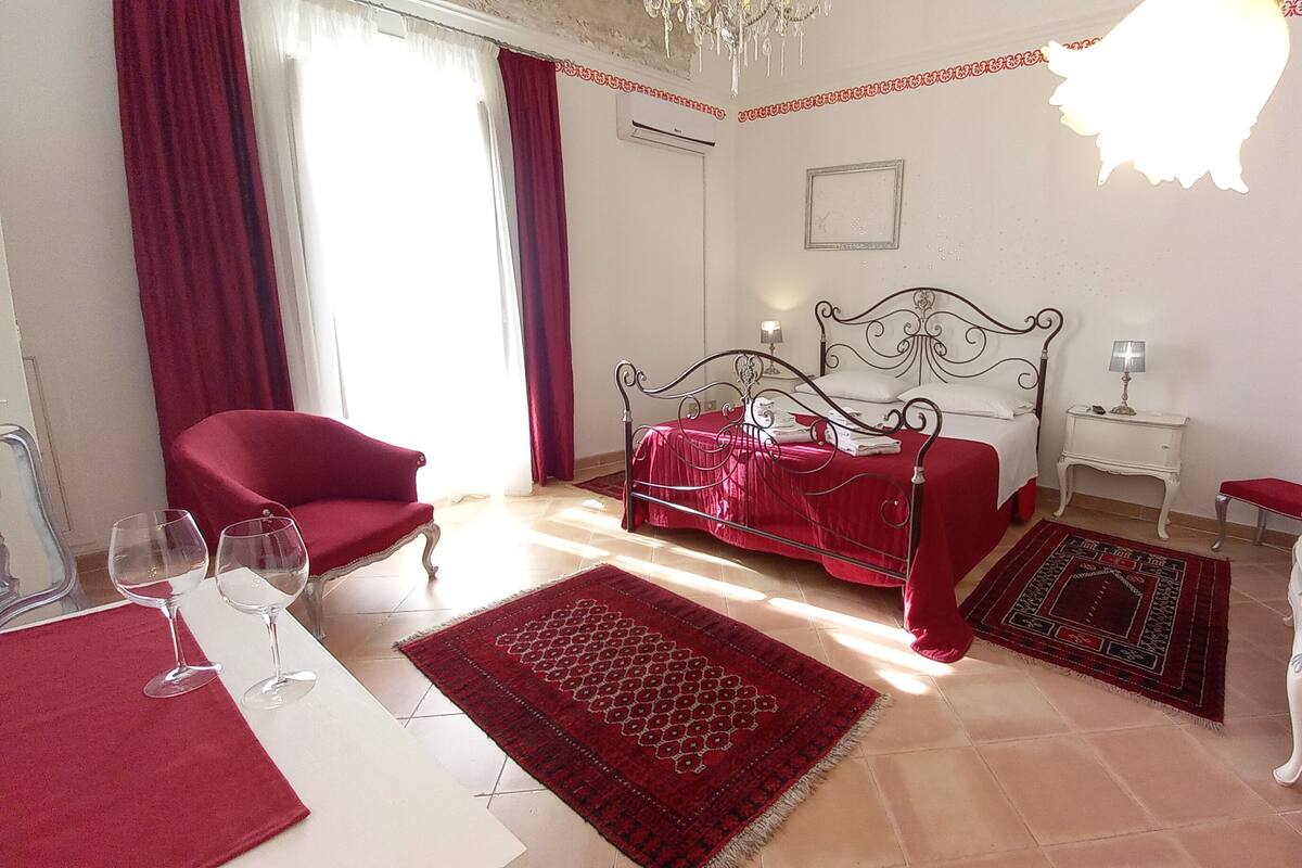 A bedroom with a bed, a red armchair, a small table with a lamp, a vase with flowers, and a chandelier.