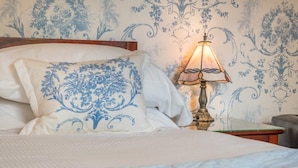 Cama com roupa de cama branca e almofadas florais azuis, mesa de cabeceira com luminária e papel de parede estampado.