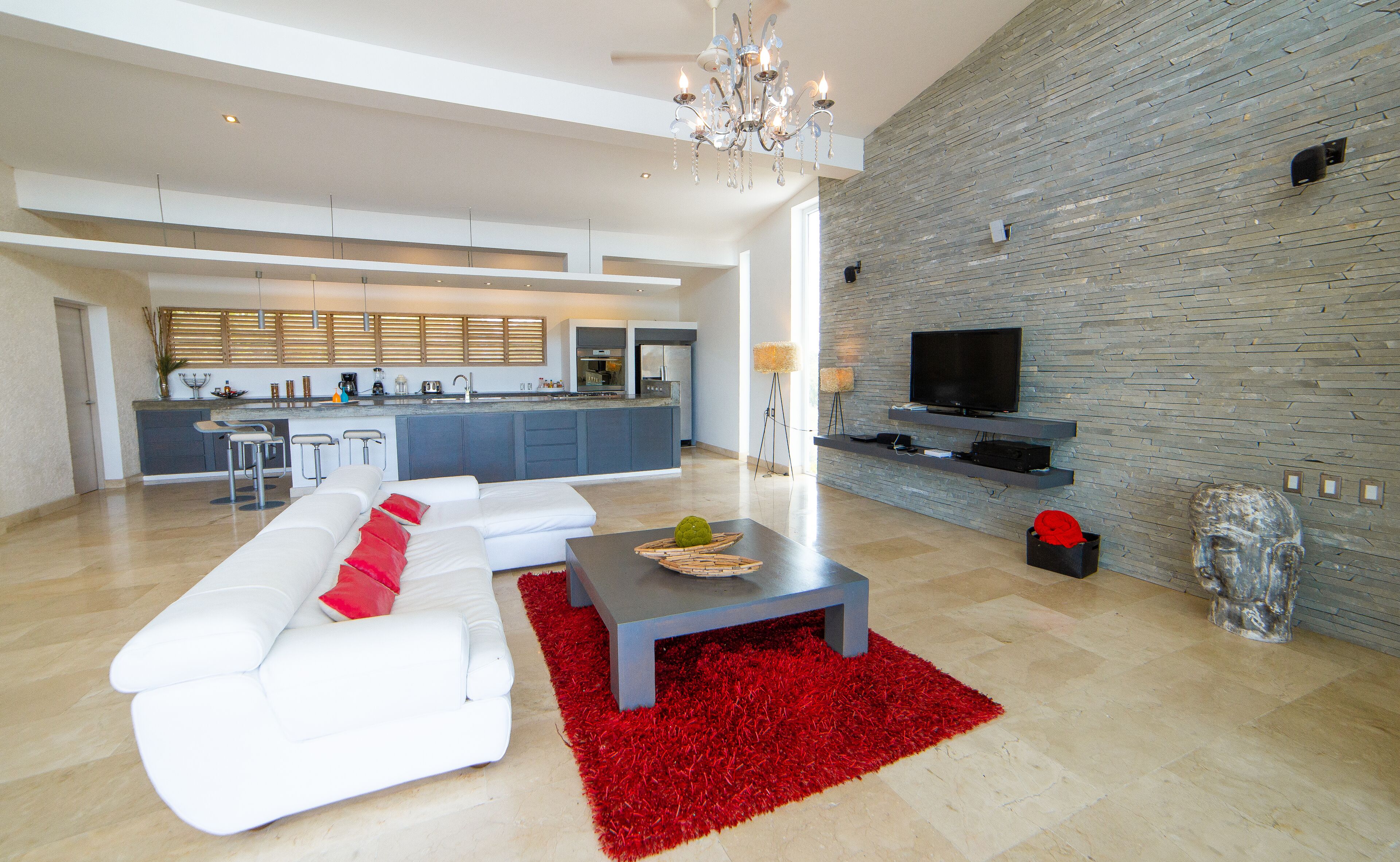 A modern living room with a white sofa, red cushions, a red rug, and a dining area with a kitchen in the background.