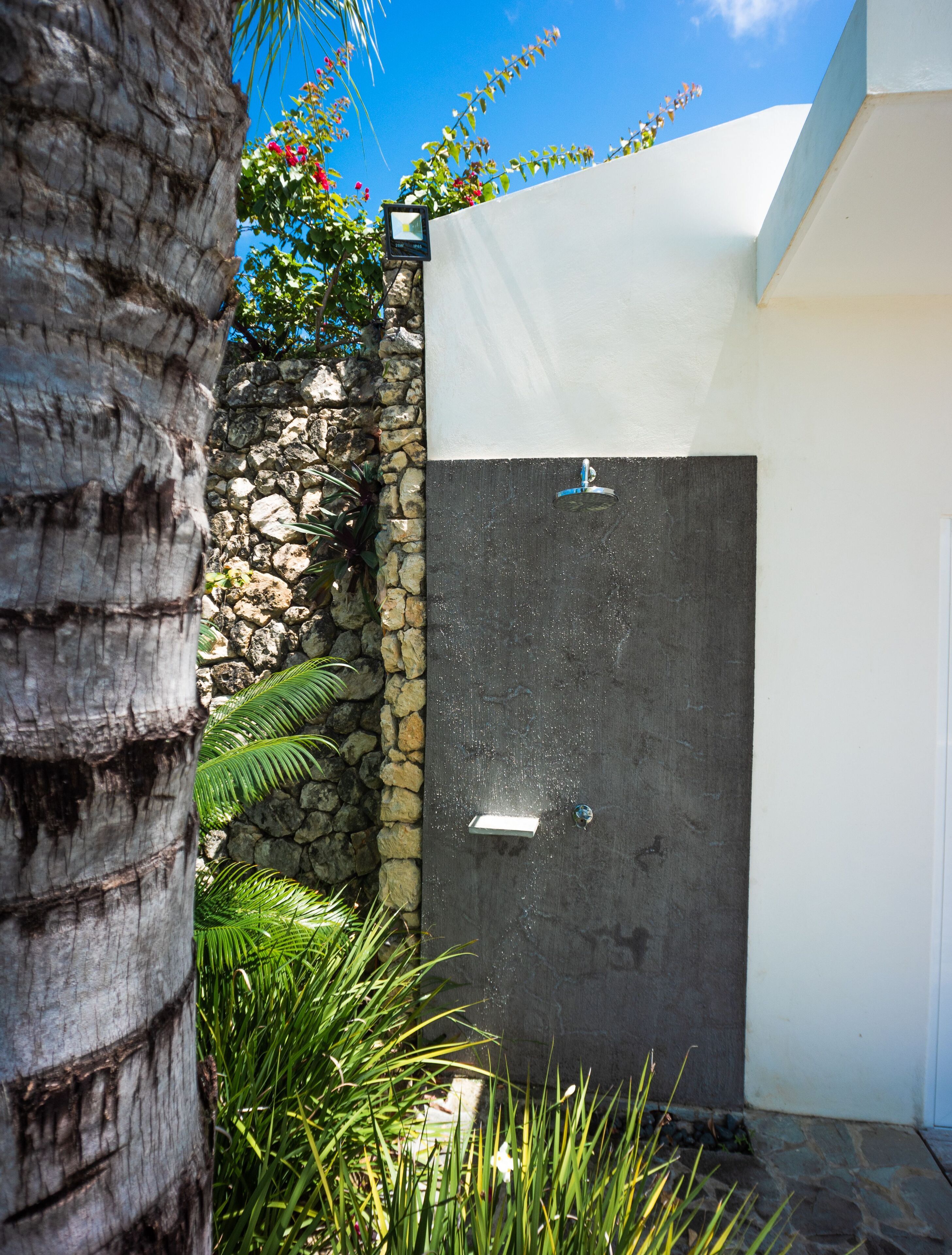 A modern outdoor shower area with a stone wall, a palm tree, and a white building in the background.