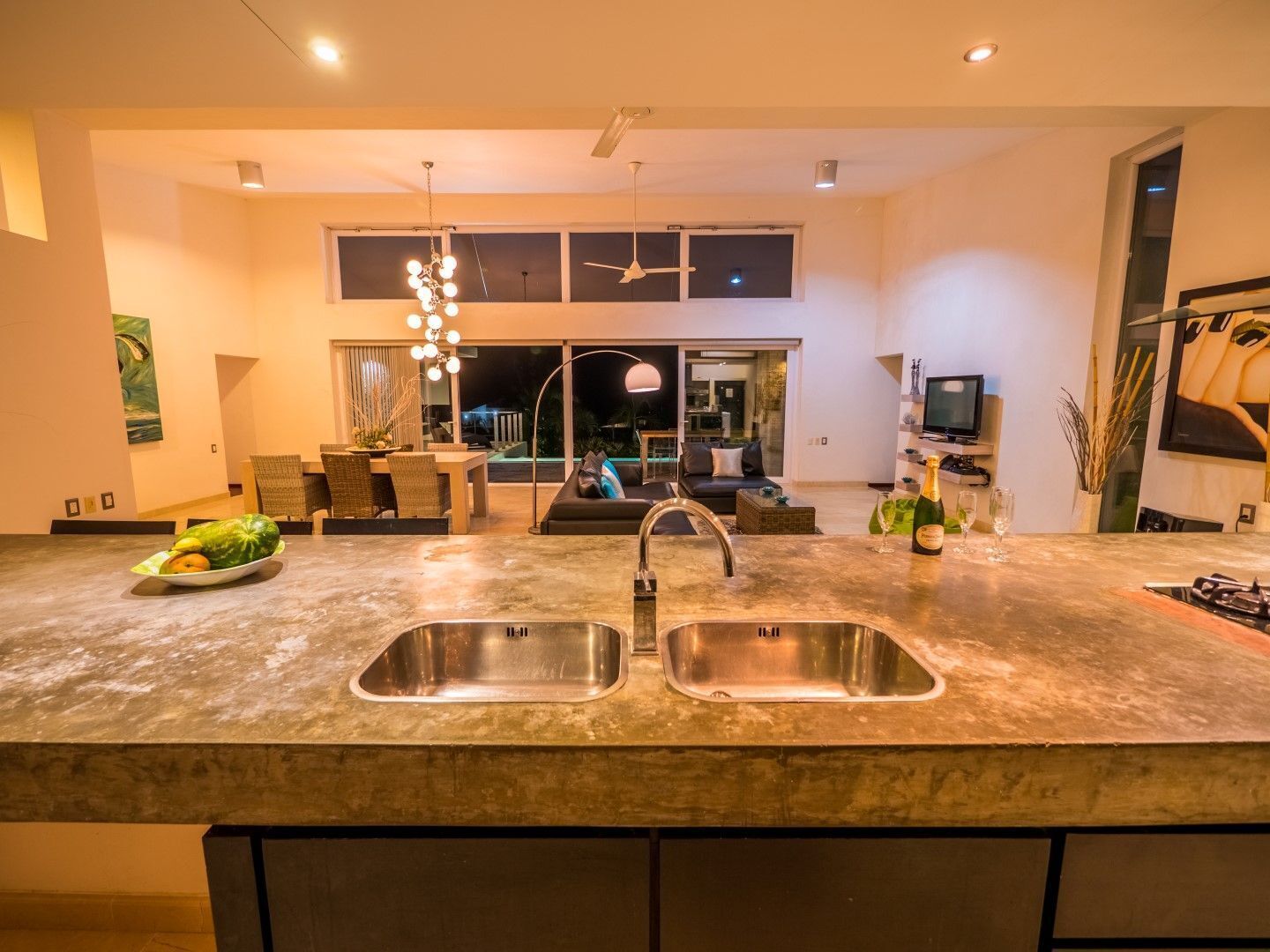 A modern kitchen with two stainless steel sinks, a dining area with a table and chairs, and a living room with a sofa and a TV.