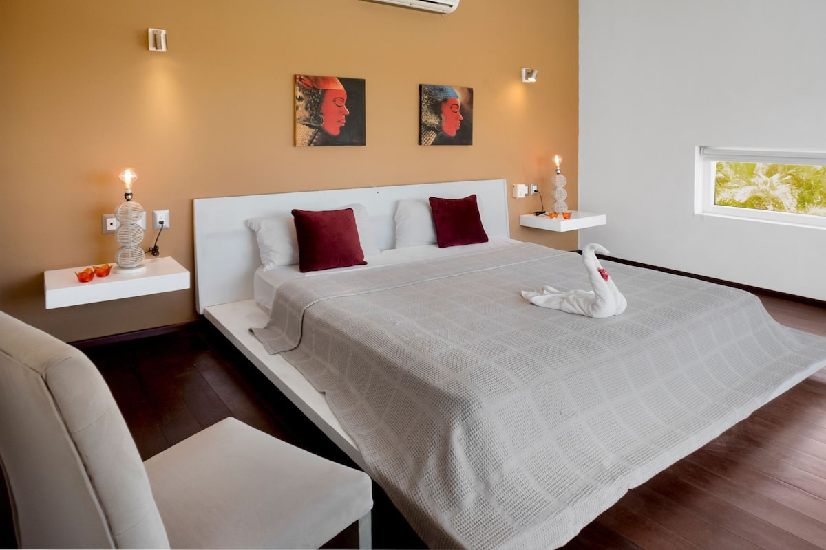 Une chambre moderne avec un grand lit, un fauteuil, une fenêtre donnant sur un beau paysage et des œuvres d’art accrochées au mur.