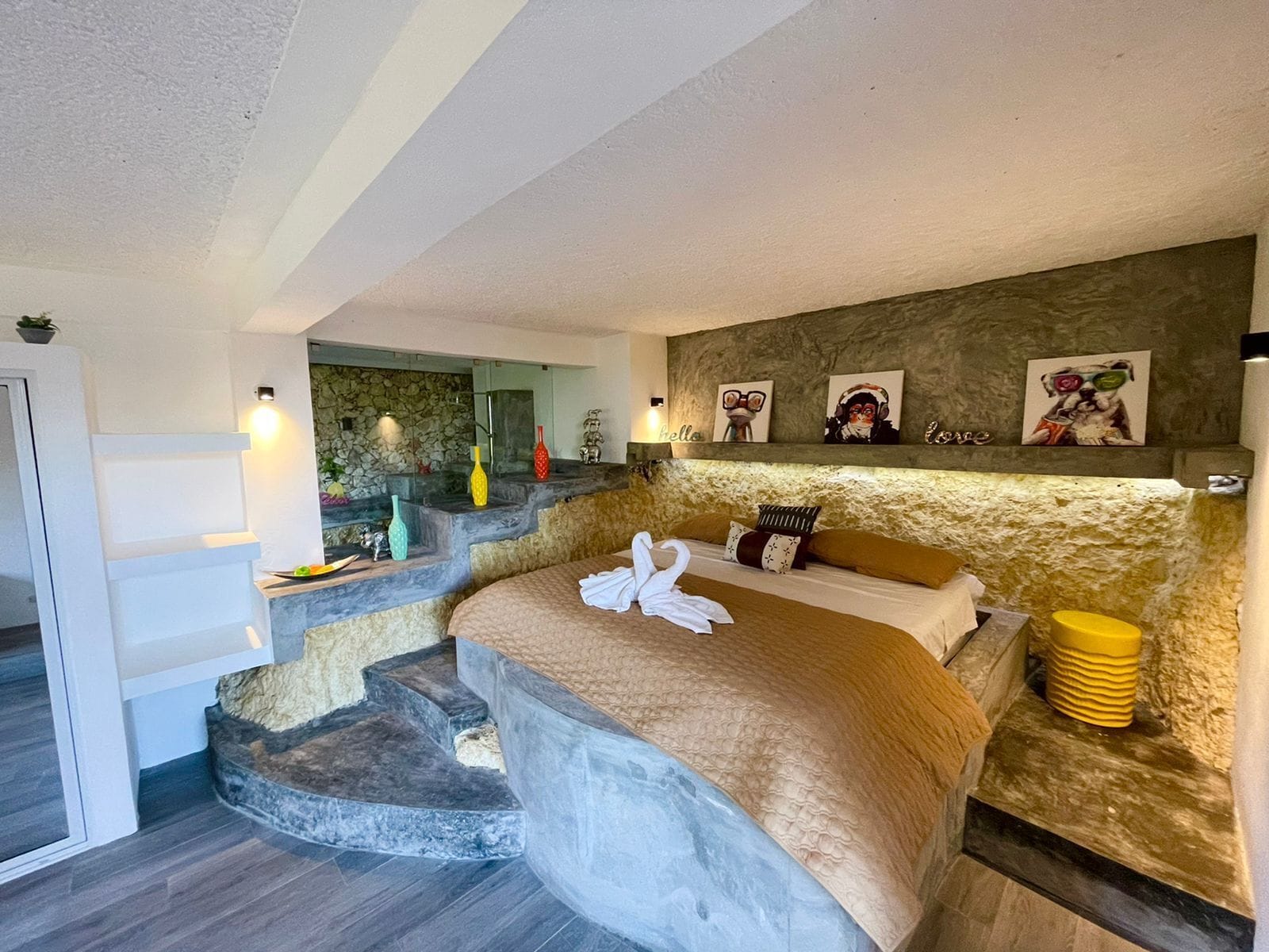 A bedroom with a stone wall, a bed with a beige blanket, two pillows, and a towel. There are two wall-mounted lights, a small shelf with decorative items, and a staircase leading to another room.