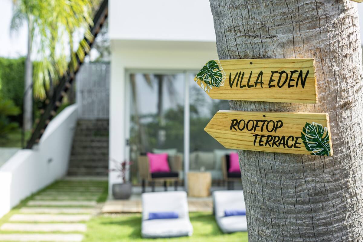 Des panneaux en bois fixés à un arbre indiquent « Villa Eden » et « Terrasse sur le toit ».