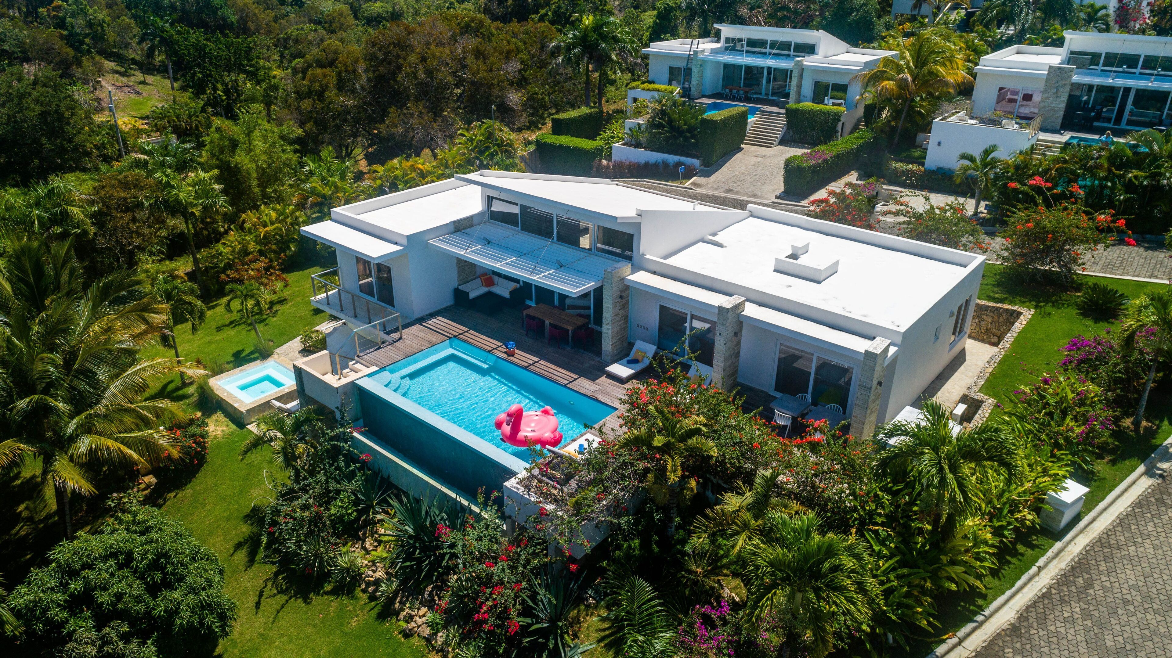 A modern two-story house with a large swimming pool, surrounded by lush greenery and palm trees.