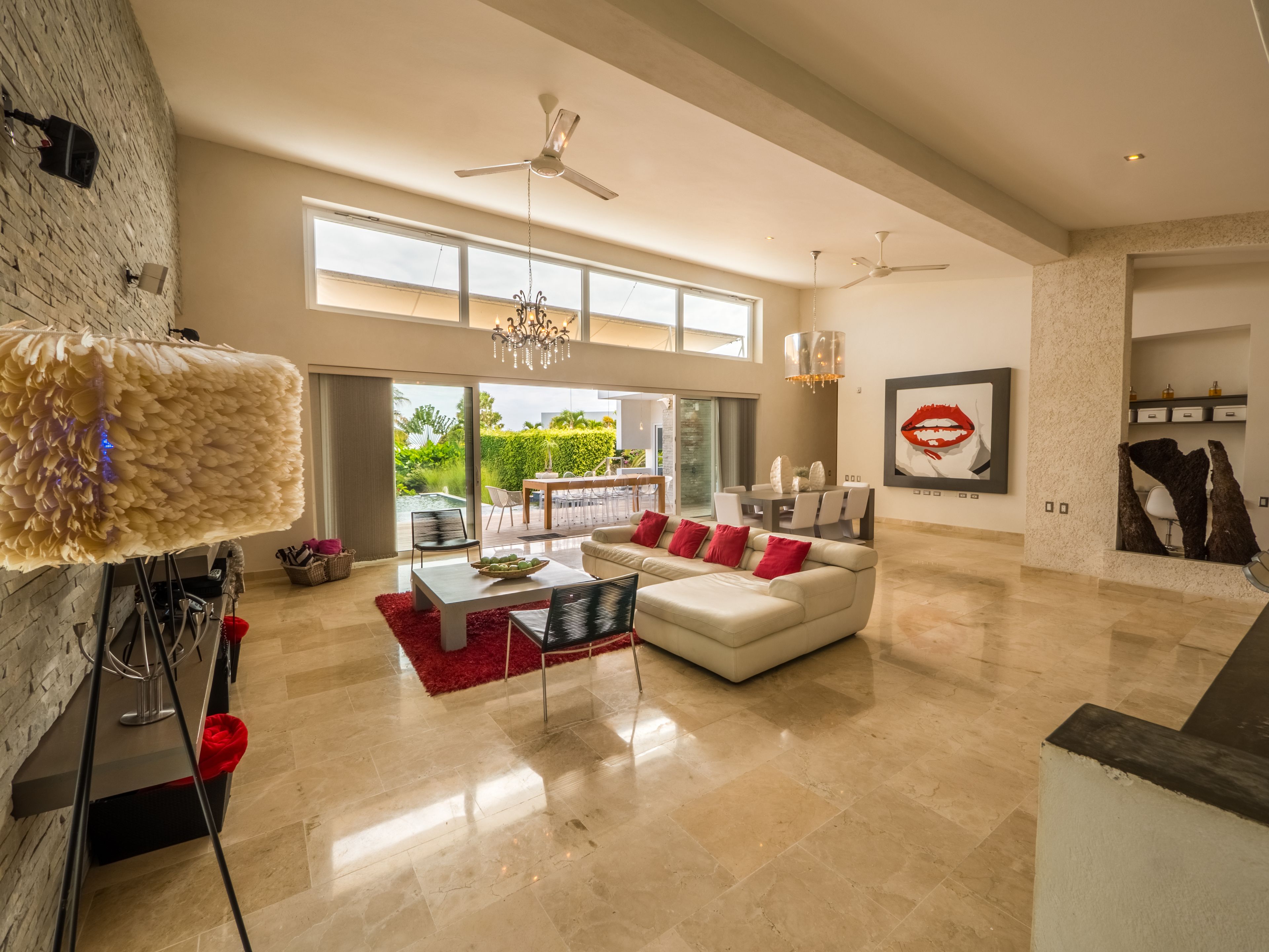 A spacious living room with a white sofa, red cushions, a coffee table, and a chandelier. There are ceiling fans, large windows, and a view of a pool and greenery outside.