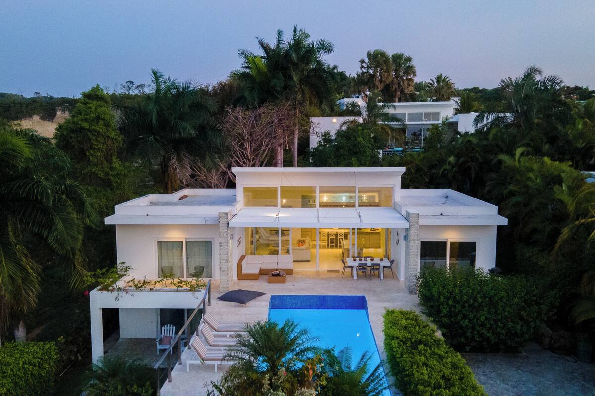Une maison moderne à deux étages avec une piscine, entourée d’une végétation luxuriante.