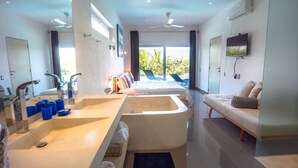A modern hotel room with a large bathtub, a sofa, a TV, and a balcony with a view of greenery. - Villas Agua Dulce (Cabarete)