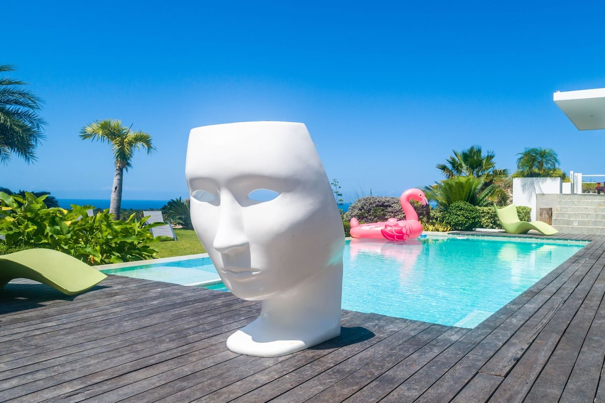 Un espace piscine agrémenté d’une sculpture blanche, d’un flotteur en forme de flamant rose et d’un bâtiment moderne offrant une vue sur l’océan.