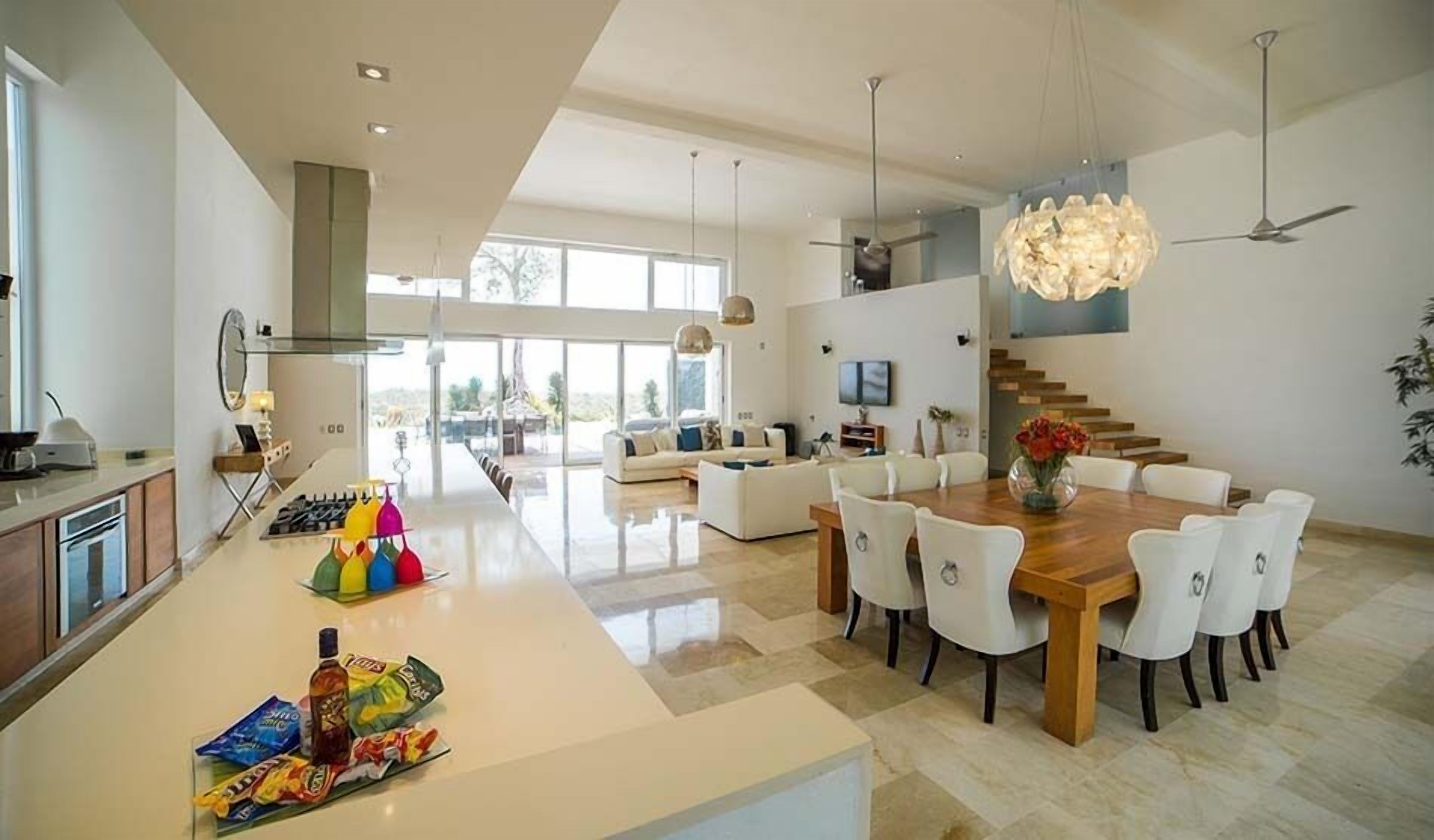 A modern kitchen with a central island, dining area with white chairs, and a staircase leading to an upper level.