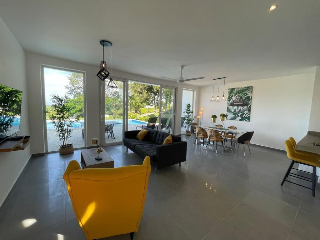 A spacious living area with a sofa, yellow chairs, a dining table, and a view of the ocean through large windows.