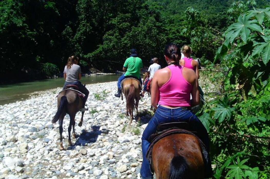 A group of people riding horses along a rocky riverbank surrounded by dense greenery.
