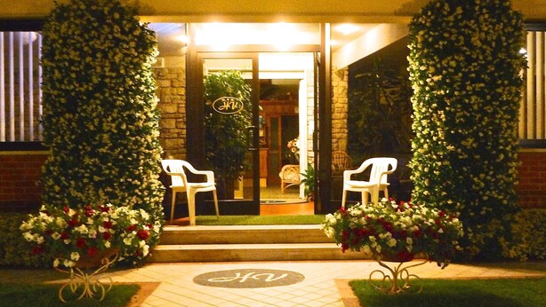 A hotel entrance with a glass door, two white plastic chairs outside, and a well-maintained garden with flowers. - Hotel Versilia (Camaiore)