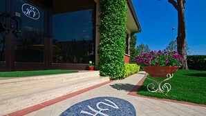 A hotel entrance with a glass door, steps leading up to it, a blue tiled area with a logo, and a planter with flowers. - Hotel Versilia (Camaiore)