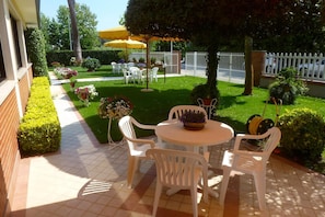 A patio with white plastic chairs and a table, surrounded by well-maintained hedges and potted plants.
