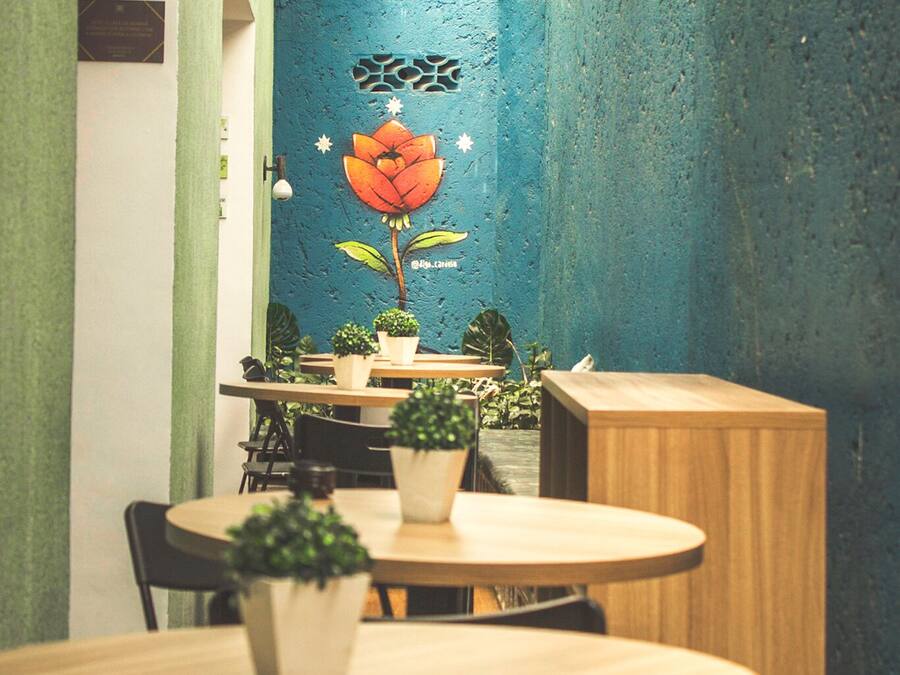 A cozy indoor dining area with round tables and chairs, potted plants, and a blue textured wall with a floral mural.