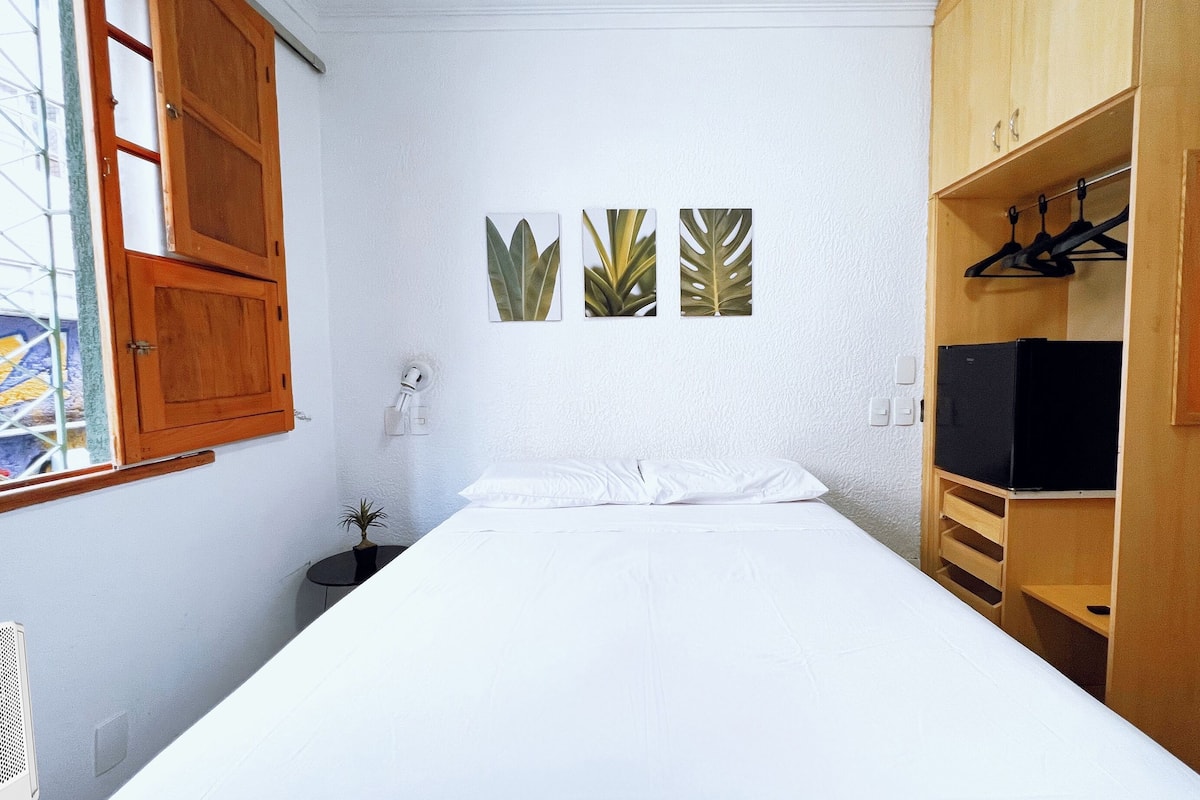 A small, neatly arranged bedroom with a single bed, a wooden wardrobe, and a window with wooden shutters.