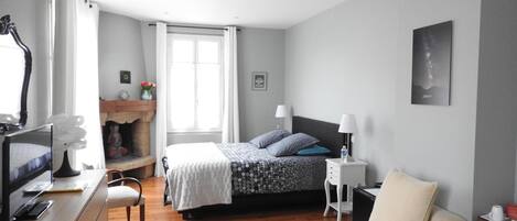 Un dormitorio con cama, mesitas de noche, televisor, sillón, sofá, chimenea y ventana.