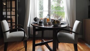 A dining area with a table set for two, a window with curtains, and a view of greenery outside. - Chambre d'hôte La Bacotterie (Bois-le-Roi)