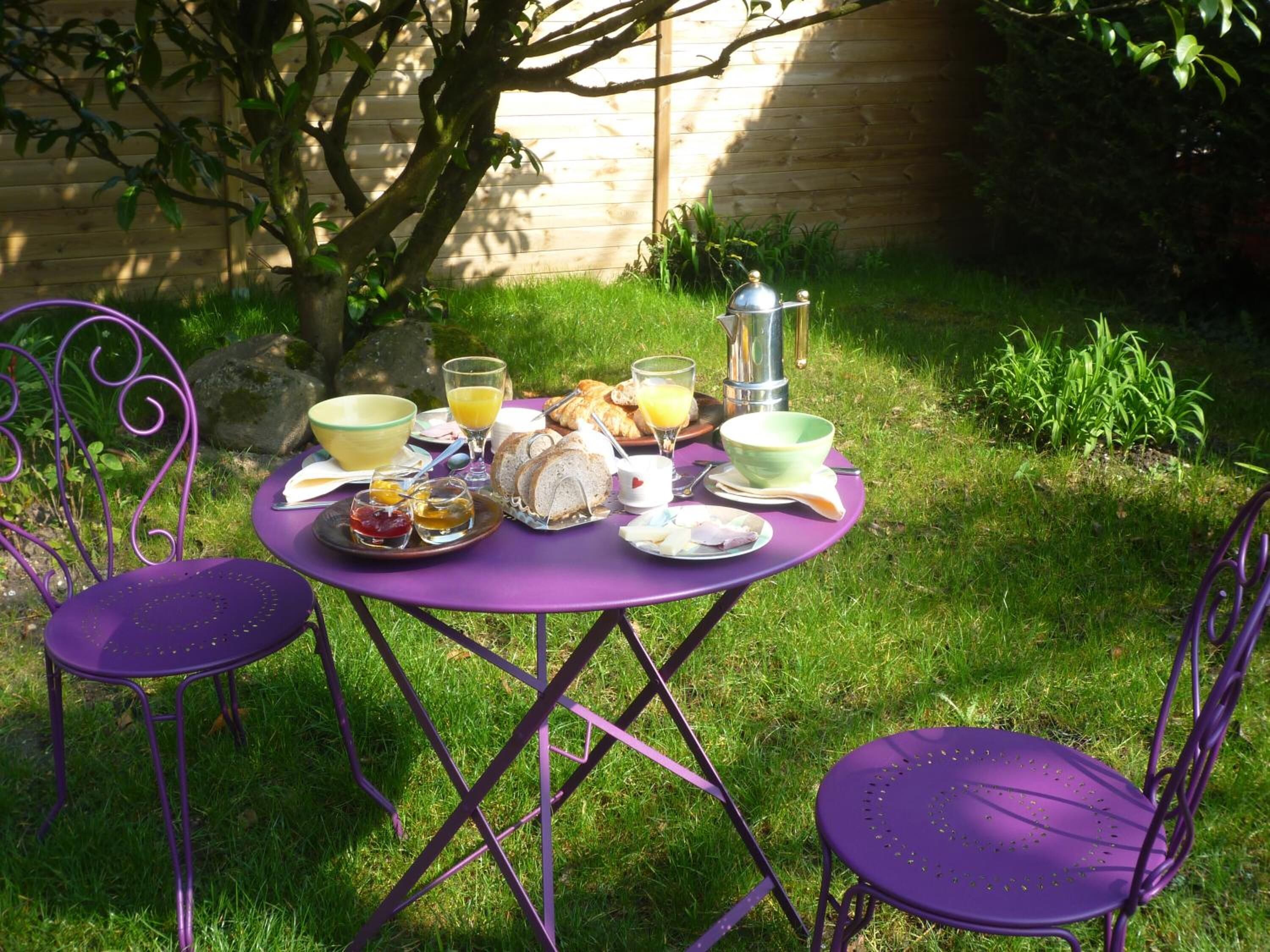 A round table with purple metal chairs set outdoors. The table is surrounded by a variety of breakfast items including bread, eggs, and a coffee pot.