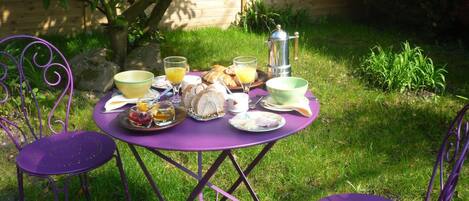 Una mesa redonda con sillas de metal moradas, colocada al aire libre. La mesa está rodeada de diversos productos para el desayuno, como pan, huevos y una cafetera.