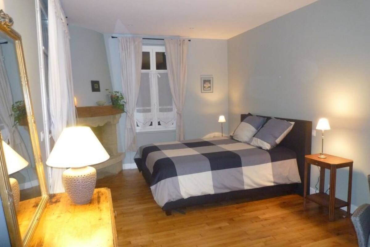 Un dormitorio con colcha a cuadros, lámpara de noche, espejo y ventana con cortinas.