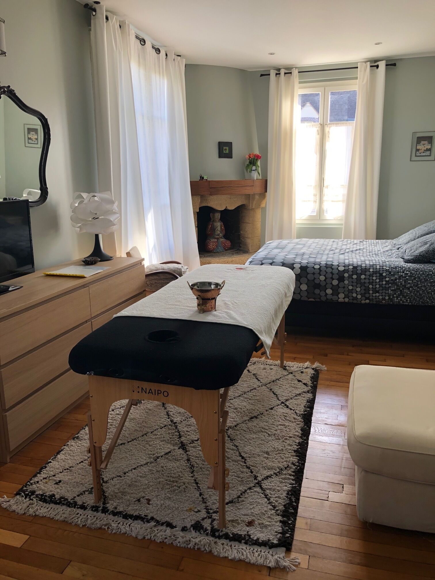 A bedroom with a wooden dresser, a black bench, a bed with patterned bedding, a white chair, and a fireplace with a Buddha statue.