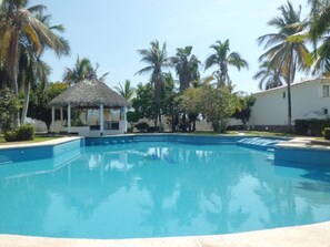 A swimming pool with a thatched-roof structure nearby, surrounded by palm trees and a white building. - Casa - Condo (Mazatlán)
