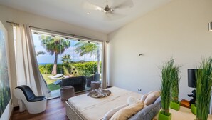 A modern bedroom with a large bed, a view of the outdoors, a ceiling fan, and a small table with decorative plants. - Villas Agua Dulce - Villa Eden (Sosúa)
