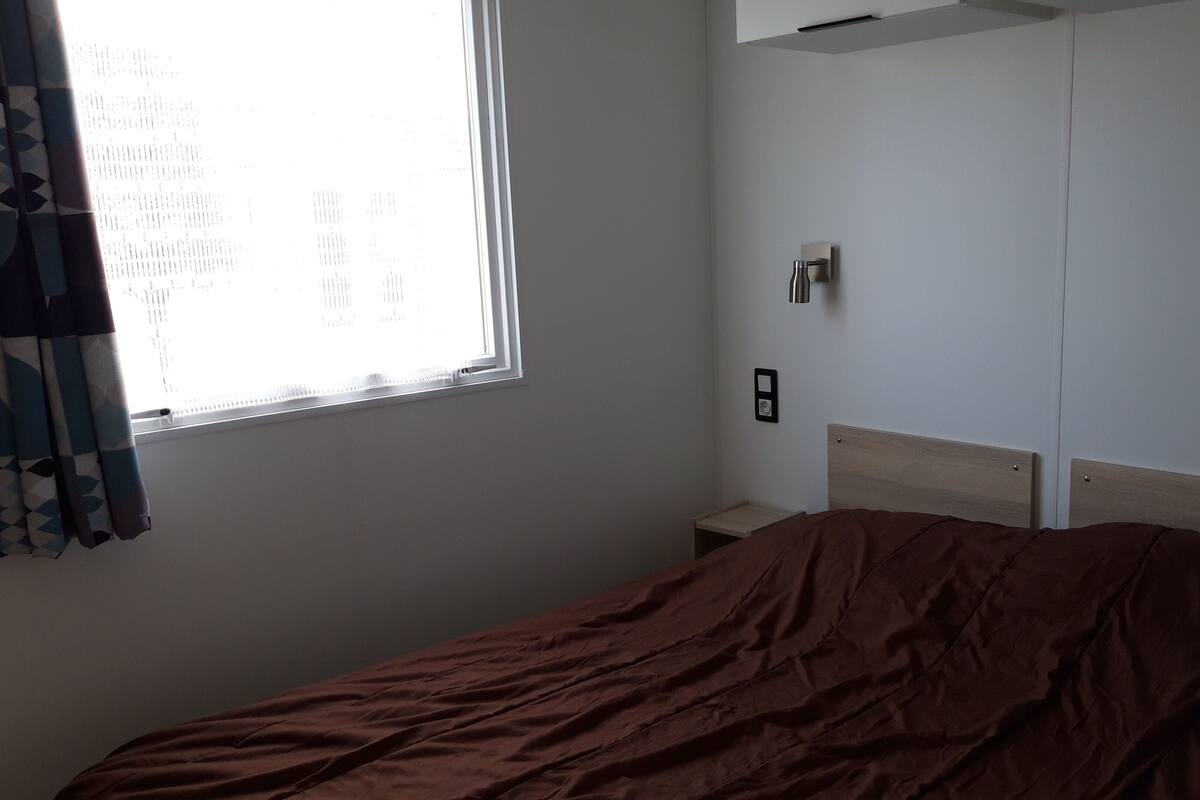 A bedroom with a bed, a window with curtains, a wall-mounted light, and a small bedside table.