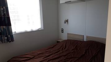 A bedroom with a bed, a window with curtains, a wall-mounted light, and a small bedside table.